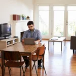 homme assis à la table à manger dans son salon étudiant son éligibilité fibre et les offres existantes sur son ordinateur portable